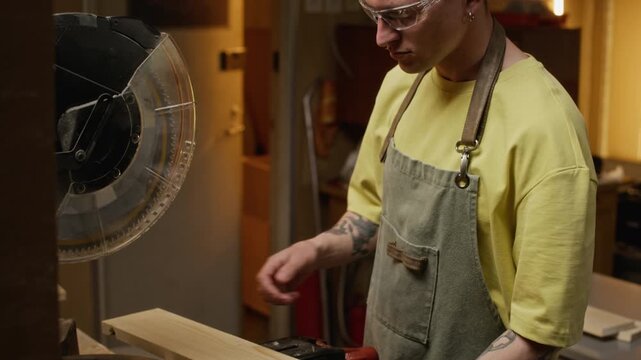 Caucasian young adult male carpenter marking wooden board with pencil and cutting it with miter saw in workshop. Video is perfect for promoting woodworking workshops or DIY tutorials