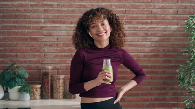 Video of sporty woman with detox juice looking at camera while listening music with earphones in the kitchen at home.
