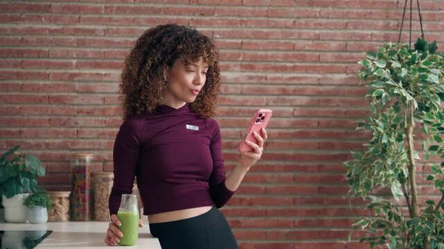 Video of sporty woman drinking handmade green smoothie while using smartphone in the kitchen at home.