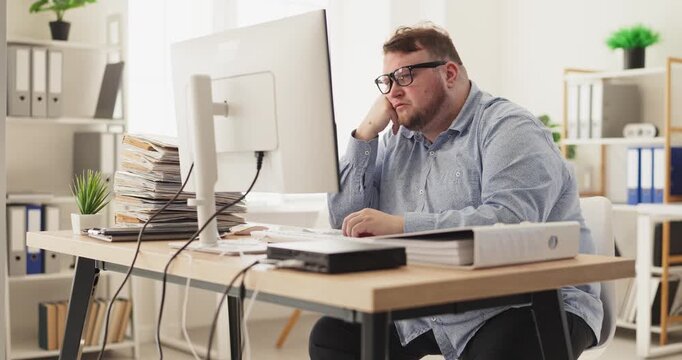 Overweight man in office faces overworked stress. A tired employee at a computer beside stacks of paperwork, showing boredom, low focus and piling tasks. Symbol of burnout and overwork.