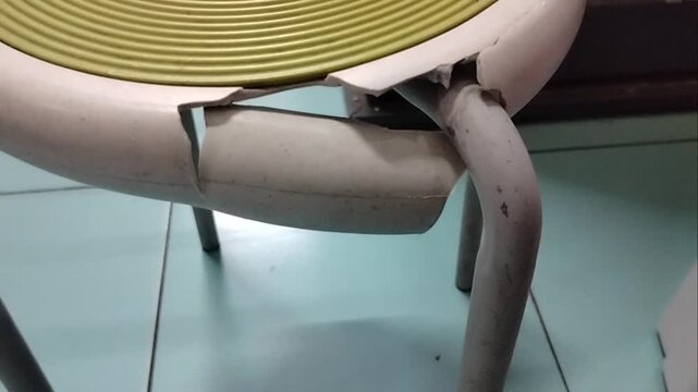 Close-up of a broken and cracked plastic round stool chair with a green textured seat cover in a room.