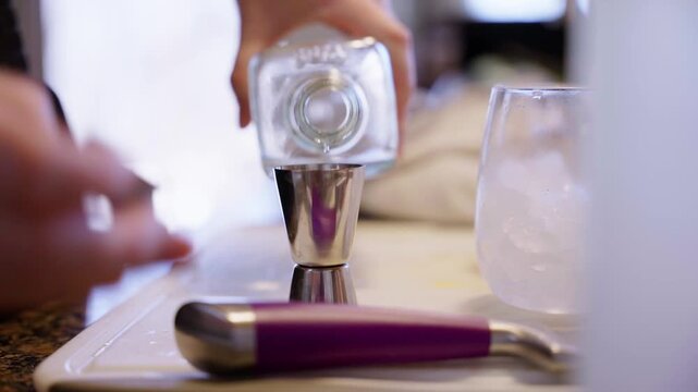 Close-up of liquor being poured from a bottle into a metal jigger beside a glass with ice, capturing cocktail preparation in a bar setting.
