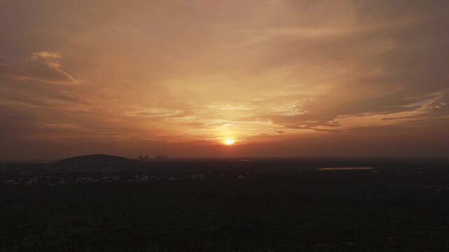 Aerial drone shot of sunset over hills and expansive landscape beneath dramatic evening sky with glowing clouds and distant waterbody during golden hour in Theni, Tamil Nadu, India