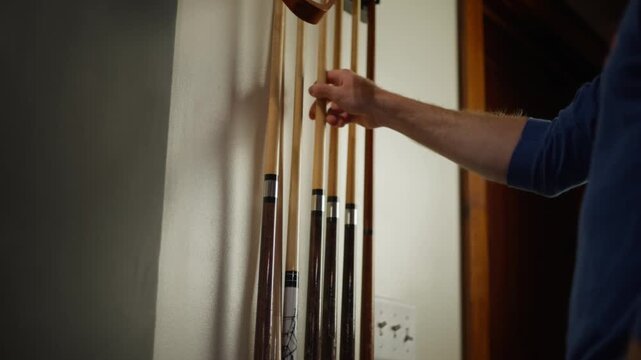 Close-up of a person selecting billiard cues from a wall rack indoors, showing wooden pool sticks, hand detail and casual game room atmosphere.
