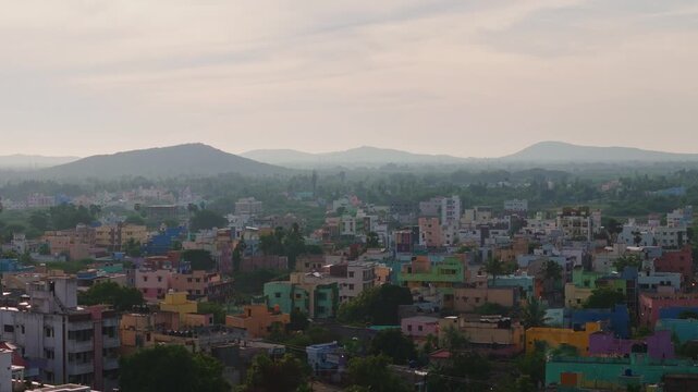 Aerial drone shot of town skyline with colorful residential buildings, lush greenery and distant mountain range beneath sunset sky in Theni, Tamil Nadu, India.