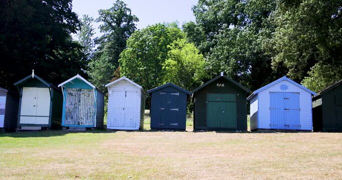 Slow motion footage of traditional colourful beach huts at Appley Beach in Ryde on the Isle of Wight with a row of seaside huts set beside mature trees on a warm summer day beneath a clear blue sky