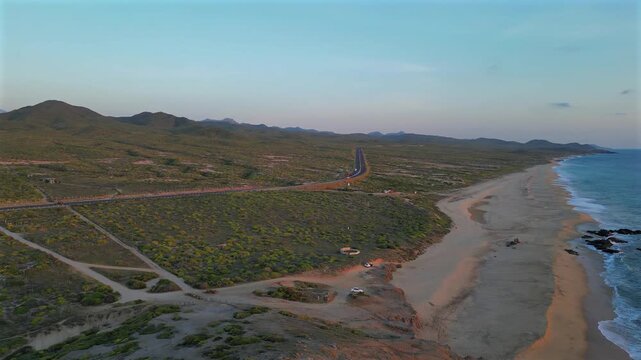 Drone rises high above Secret Rocks Beach below Todos Santos, overlooking a rare lush green desert and distant mountain range with cars passing along a highway below, before turning to reveal the sout