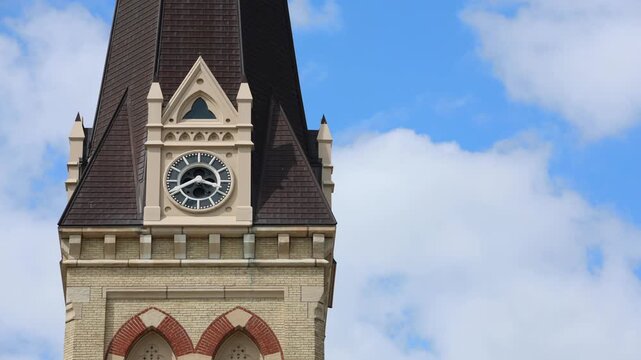 A timelapse video still showcasing an old architectural church clock tower. The clock hands move as white clouds quickly pass across a blue sky in the background during daytime.