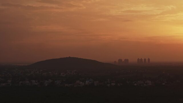 Aerial drone shot of sunset casting warm golden light over hill, town skyline and distant high rise buildings beneath hazy sky in Theni, Tamil Nadu, India