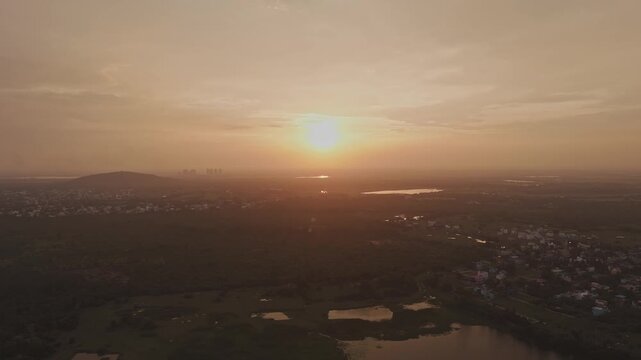 Aerial drone shot of glowing sunset over hills, forests and expansive landscape beneath golden sky with distant waterbodies reflecting warm light in Theni, Tamil Nadu, India