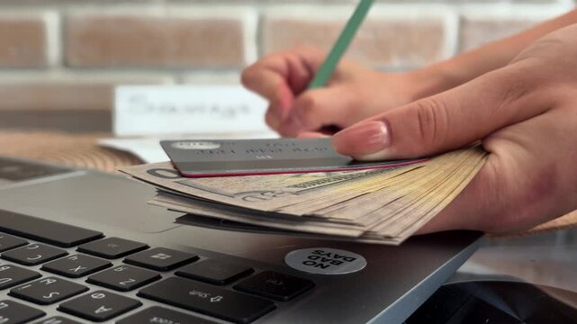 Woman's hands managing personal finances by writing down savings while holding a stack of dollar bills and a credit card next to a laptop on a desk in an office