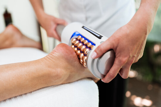 A professional therapist performs a body contouring massage using an innovative roller device for cellulite reduction and lymphatic drainage on a client's leg and foot.