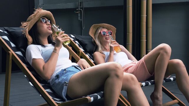 Two female friends wear straw hats and sunglasses relax on deck chairs, drink cold beverages laugh together on sunny day. Summer vacation, friendship, leisure, and carefree poolside lifestyle