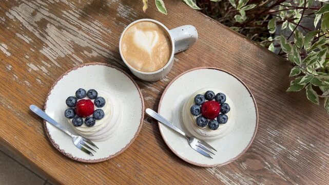 Top view of two mini Pavlova desserts with fresh blueberries and strawberries and cappuccino on a rustic wooden table at a cozy outdoor caf&eacute;.