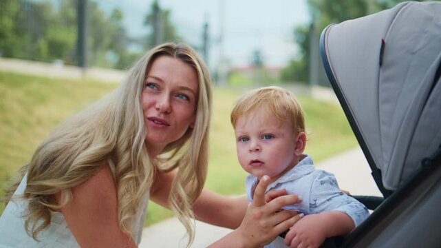 Blonde woman smiles as she helps a toddler into a stroller. She holds the child's arm gently. The child looks up curiously at the camera. Green grass and trees form a park backdrop