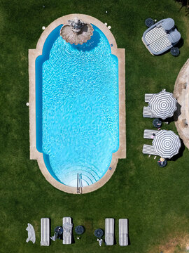 Aerial view of an ornate swimming pool with a stone fountain, surrounded by a lush green lawn with striped lounge chairs and umbrellas in Cascais, Porto, Portugal.