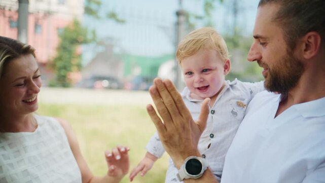 Young couple plays with baby outdoors. Mother claps hands, smiling joyfully. Father holds baby, also clapping, looking happy. Baby looks at camera, curious and calm