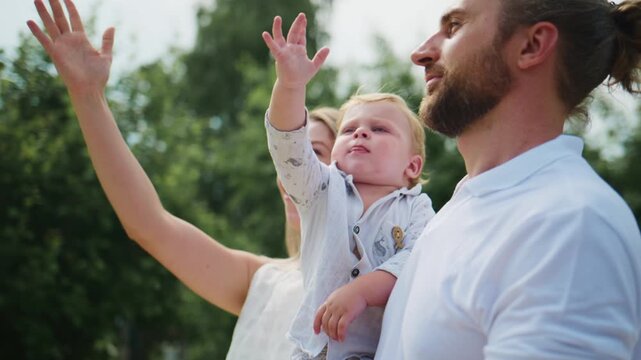 Happy family outdoors, baby held by father, mother waving. Parents smile warmly, child looks curious. Scene captures joyful family moment in park. Ideal for parenting, family bonding