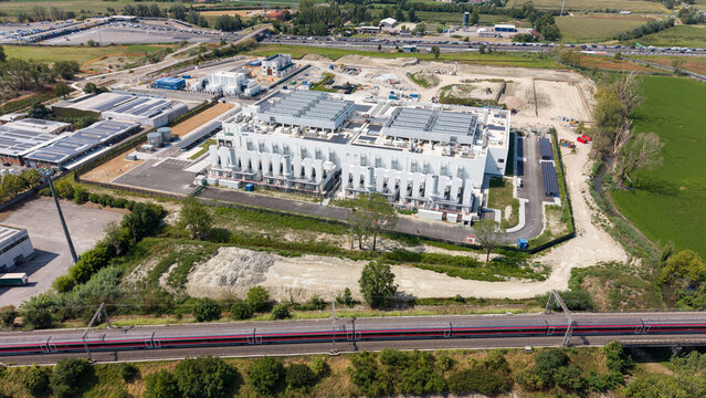 Aerial view of a modern data center facility featuring advanced cooling systems and digital infrastructure surrounded by green fields and a railway line in Melegnano, Lombardy, Italy.