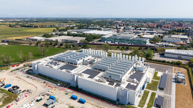 Aerial view of a large modern data center facility featuring extensive rooftop cooling units and solar panels, surrounded by industrial infrastructure in Melegnano, Lombardy, Italy.