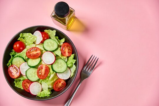 Fresh garden salad with lettuce tomatoes cucumber and radish in dark bowl on pastel pink background with fork