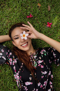 Smiling model lying on green grass holding a flower over one eye