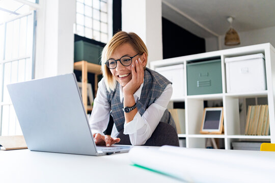 Smiling businesswoman with hand on chin using laptop while leaning on desk