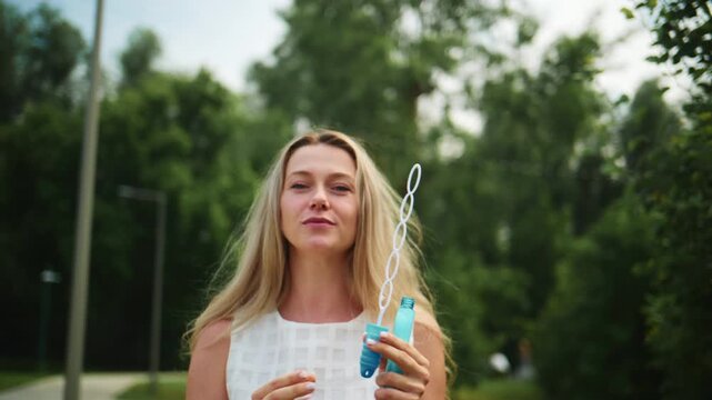 Young woman blows bubbles outdoors with a wand. She smiles happily, eyes focused on floating spheres. Green trees and open park space surround her. Footage captures joyful summer moments