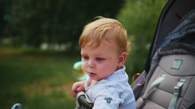 Curious toddler in a stroller watches colorful bubbles float by. The child's wide eyes show wonder and delight as they reach toward the floating spheres. Soft, natural light bathes the scene
