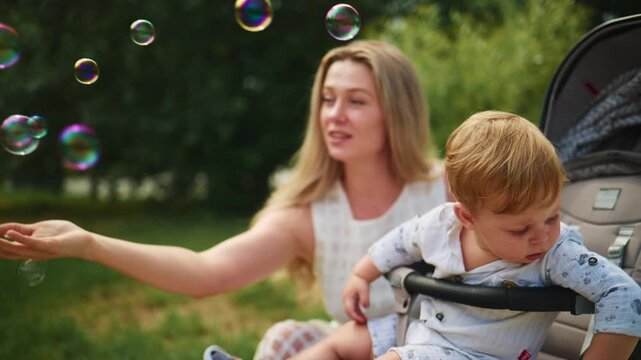 Mother smiles while blowing bubbles near baby in stroller. Child looks curiously at floating soap bubbles. Scene shows happy family enjoying sunny park day. Perfect for parenting, family