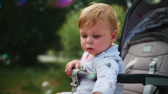 Curious toddler in a stroller gazes at floating bubbles. Sunlight glints through bubbles near the child's face. The baby's focused expression shows wonder and delight