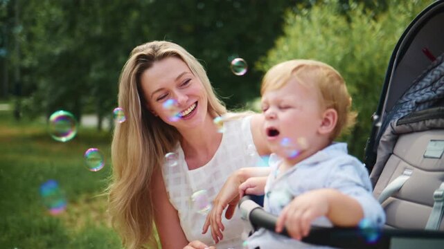 Mother smiles as baby watches floating bubbles outdoors. She leans close, eyes bright with joy. Baby reaches toward bubbles with curious hands. Scene captures tender, playful moment in sunny park
