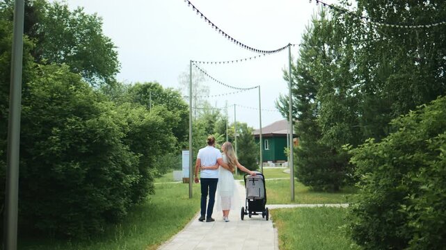 Couple walks together, strolling along tree-lined path. They push baby stroller, smiling, enjoying outdoor stroll. Greenery surrounds them, creating peaceful atmosphere