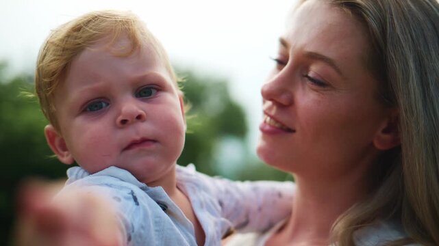 Mother holds toddler outside. Child points forward with curious eyes. Woman smiles warmly, gaze loving. Scene captures playful, bonding outdoor moment. Ideal for parenting, family, or childhood themes