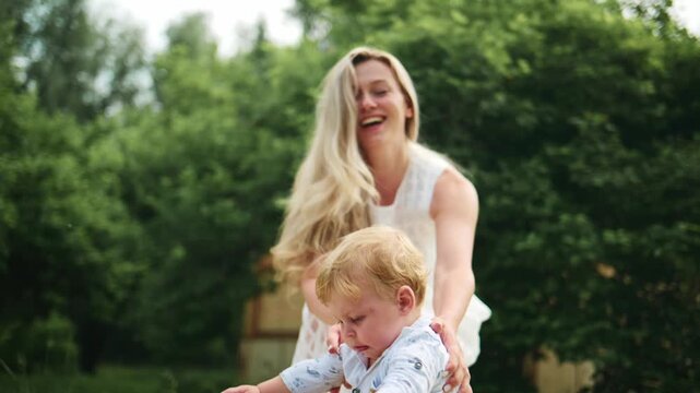 Young child walks outdoors with supportive adult. Mother smiles warmly, encouraging child's steps. Gentle summer scene with green trees. Ideal for family, parenting, or early childhood themes