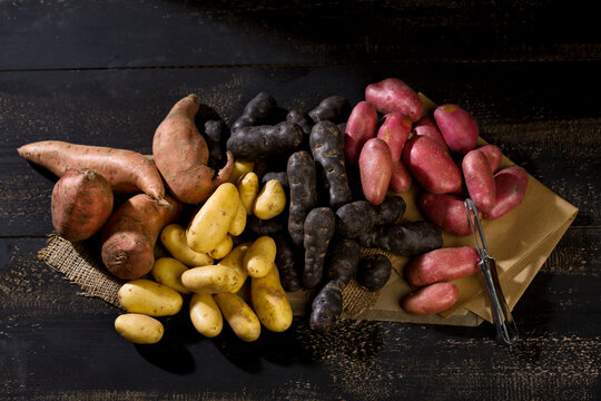 Different sorts of potatoes and peeler on brown paper, jute and dark wood