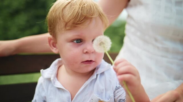 Little boy sits on bench, examining small object in hands. Mother in white dress sits beside him, partially visible. Boy looks curious and focused, exploring item with care