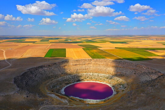 Aerial view of Meyil Lake, a sinkhole lake with vibrant pink water, surrounded by vast agricultural fields and patchwork farmland under a blue sky in Karapinar, Konya, Turkiye.