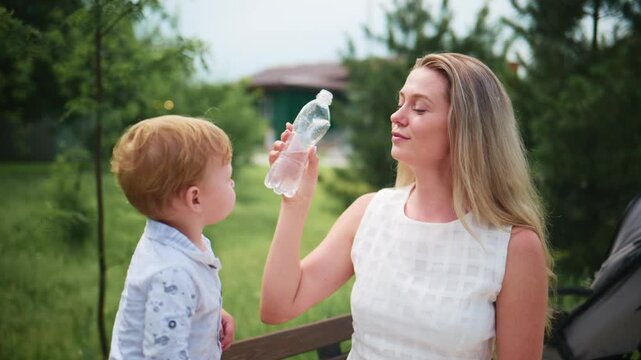 Mother and toddler drink water together outdoors. Both smile warmly as they sip from clear bottles. Scene captures peaceful, healthy bonding moment. Ideal for parenting, health, or lifestyle content