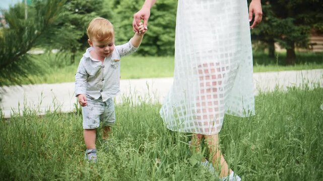 Toddler holds his mother's hand while walking through tall grass. The child looks down with curiosity. Mother walks calmly beside him in a light dress