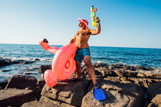 Man standing while holding flamingo ring and squirt gun against sea