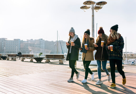 Four friends with coffee to go and cell phones walking on promenade in winter