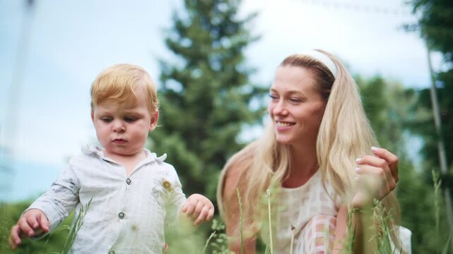 Woman and child enjoy sunny day in grass. Mother blows dandelion seeds to toddler. Child watches with curious expression. Mother smiles warmly. Scene captures playful outdoor bonding moment