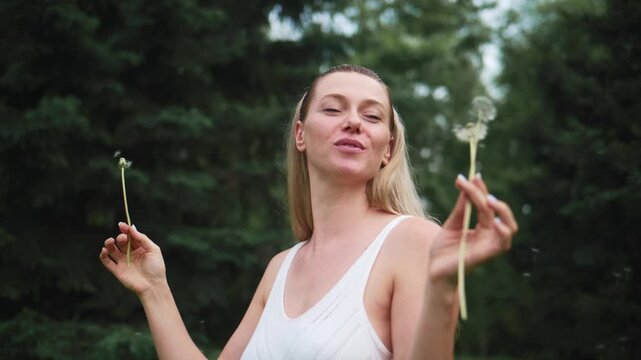Woman smiles while blowing dandelion seeds. Forest backdrop adds natural charm. Gentle breeze carries seeds into air. Perfect for summer, nature, or dreamy themes. Ideal for wellness, childhood