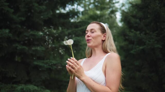 Young woman in white dress blows dandelion seeds in forest. Her eyes closed, smile shows peaceful joy. Dandelion seeds float gently into air. Soft natural light surrounds her calm moment