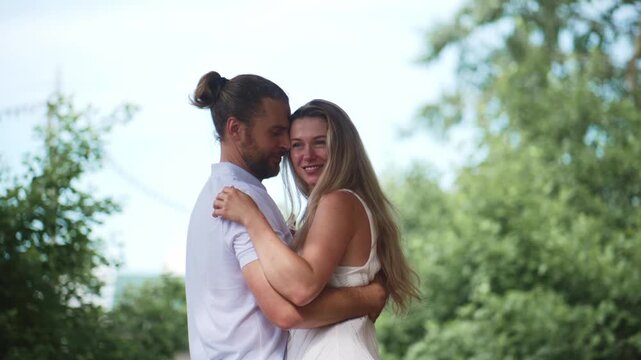Loving couple shares a tender kiss in a sunny outdoor setting. The man, with a bun hairstyle, embraces the woman gently. The woman, with long hair, smiles happily as they connect