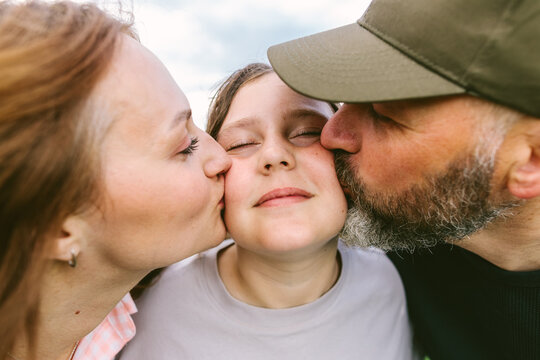 Mother and father kiss their daughter on the cheeks outdoors