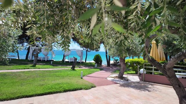 The terrace of a luxury island hotel overlooks the turquoise Aegean Sea, a green garden with olive trees, and a private beach in sunny Greece
