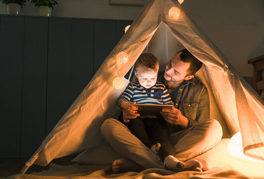 Father and son sharing a tablet in a dark tent at home