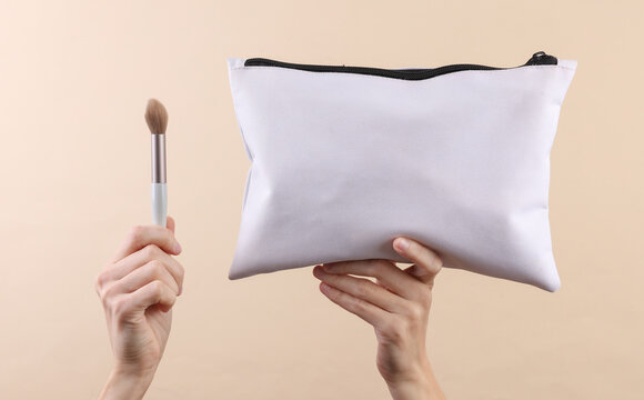 Hands holding White fabric cosmetic bag and makeup brush on a beige background. Beauty concept, mockup for design.